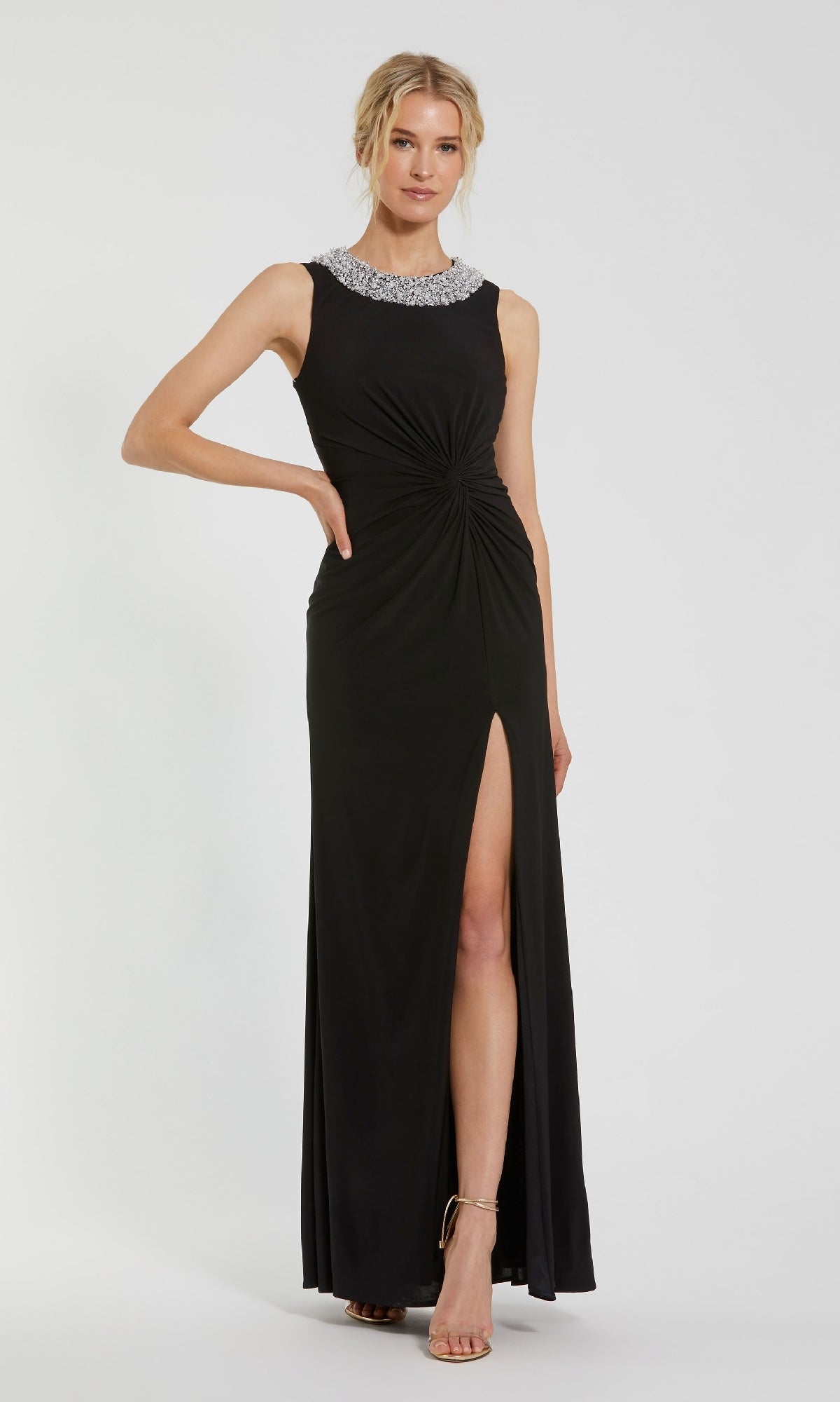 A woman in the Mac Duggal 49952 Jeweled Long Black Formal Dress, featuring a jeweled neckline, gathered waist, and thigh-high slit, pairs it with nude high-heeled sandals and stands elegantly against a plain background.